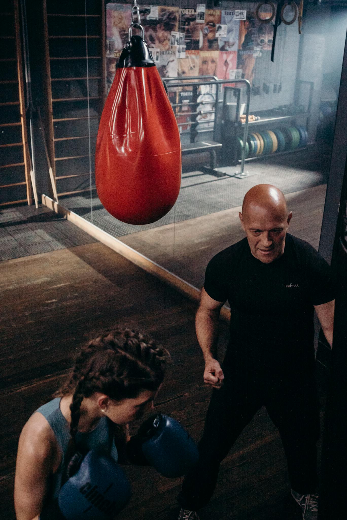 Female boxer practicing punches with coach below red punching bag in gym.