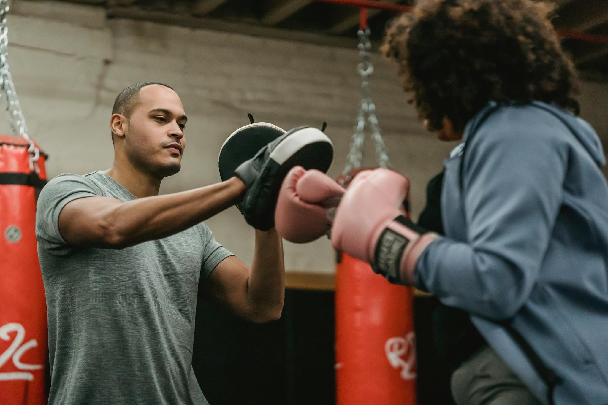 Strong African American personal instructor with boxing paws training anonymous female with gloves while practicing in gym with punching bag